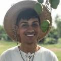 photo of a smiling Rami wearing a white shirt and straw hat in front of a mango tree branch. Rami has light brown skin with black hair and piercings between the eyes on his nose bridge and on the earlobes. Four green mangoes hang from the top-right edge of the frame and dangle around Rami’s face, covering parts of the straw hat. The background is a green open field with out of focus, tall trees.