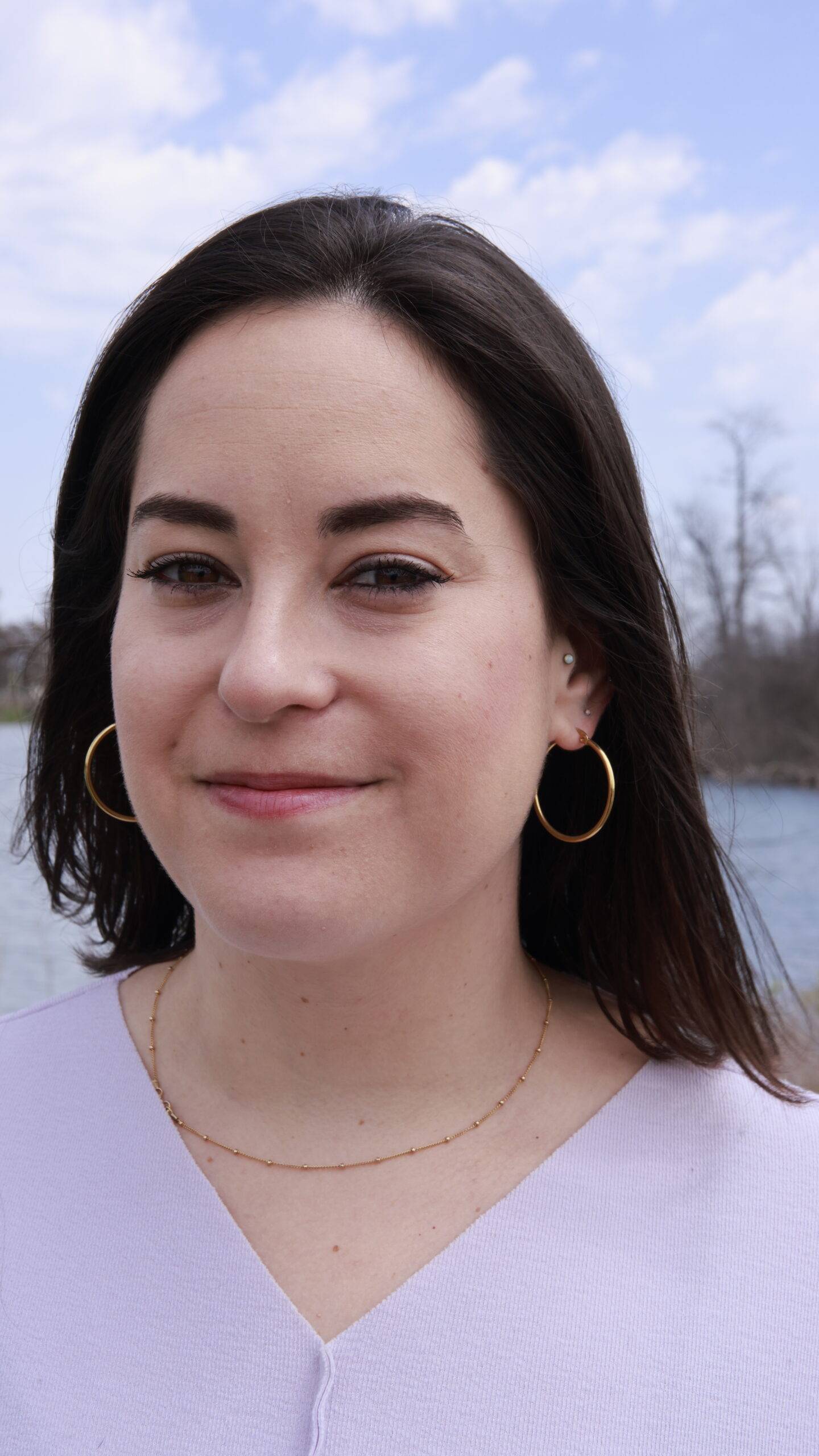 Portrait of Ani from the chest up. Her straight dark hair blows back in the wind and she smirks with a closed-mouth smile. She has white skin, and wears gold earrings, a gold chain necklace and a lavender v neck shirt that seams down the middle. 